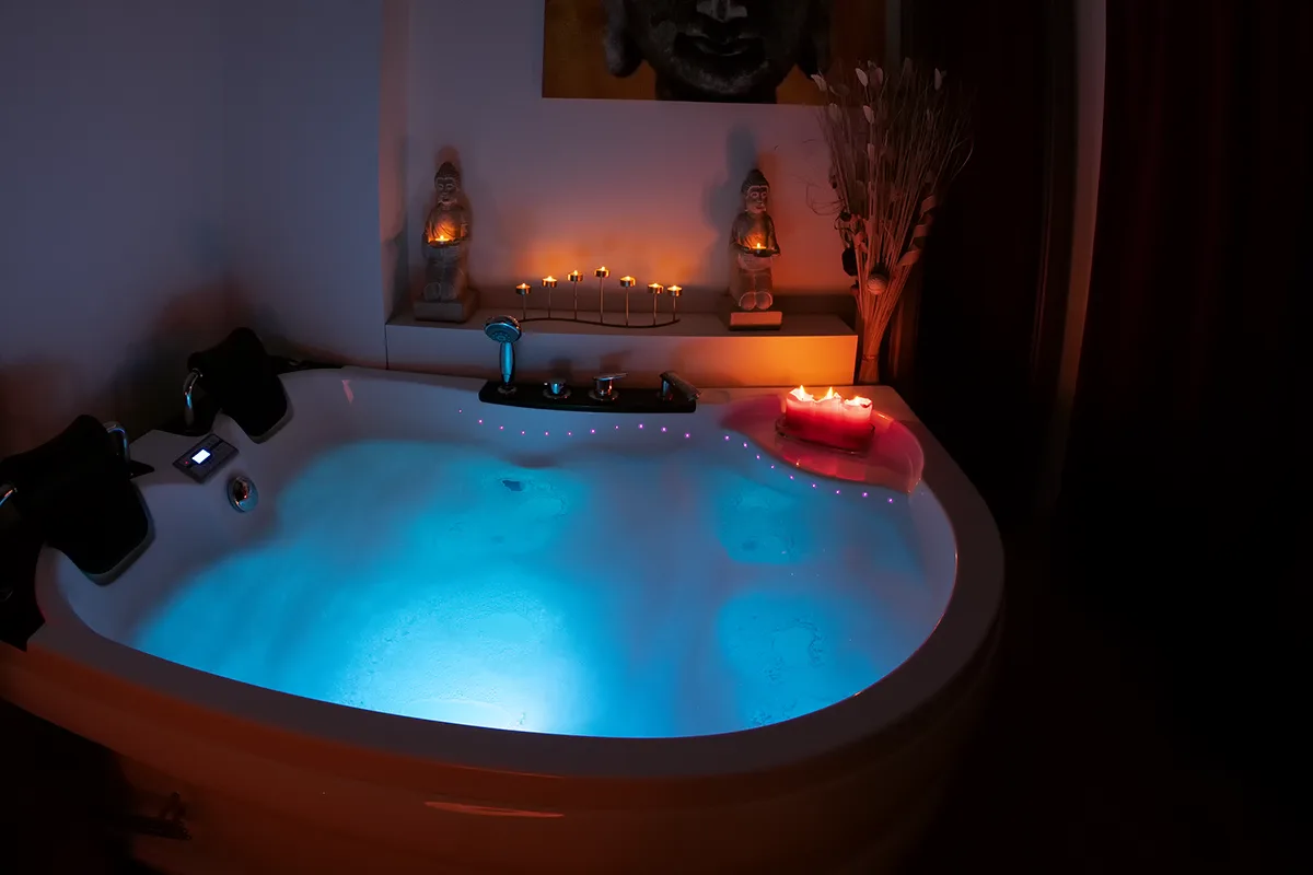 Jacuzzi de esquina con el agua iluminada en azul, velas rojas y figuras de Buda en la repisa