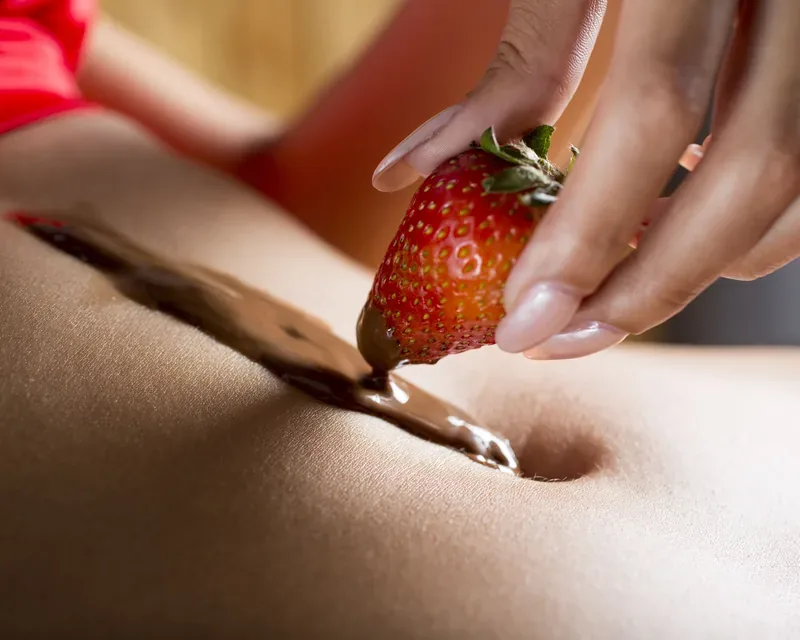 Pink-nailed hand drawing a strawberry through chocolate on skin, massage menu with two masseuses in Zaragoza